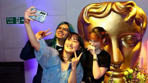 A group of three young people pose for a selfie in front of a large, golden Bafta mask statue. The young woman at the front of the group holds up her camera phone and throws up a peace sign, as does the young man with green hair to her right. A young woman to her left places the back of her hand against her mouth as she giggles.