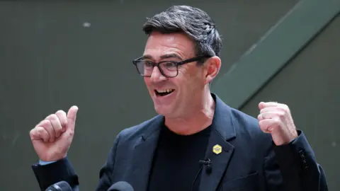 Reuters Andy Burnham with black-framed glasses, a black T-shirt and dark jacket with with both hands raised