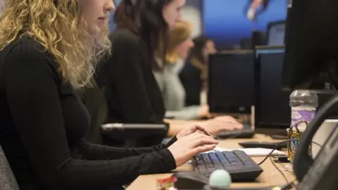 A profile view of a woman with curly blonde hair at an office desk looking at a screen with her hands hovering over a keyboard.