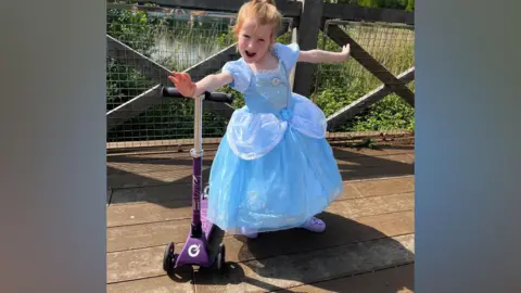 Handout A five-year-old girl in a long blue dress and purple shoes. She is standing with her arms outstretched near her scooter, at a park.
