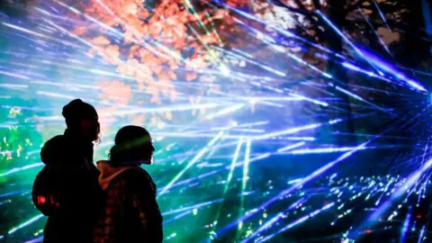 Westonbirt Arboretum The silhouettes of a woman and a girl - perhaps in her early teens - who are wrapped up warm can be seen as they look at blue and green strobe lights beneath brown autumn trees.