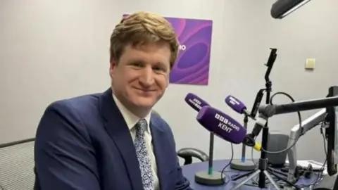 Jack Valpy/BBC A man with brown hair and wearing a blue suit sits at a desk. He's smiling at the camera and there is a purple microphone in front of him, with BBC Radio Kent branding