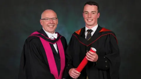 Father and son John and Cameron Gibson, pictured inside at Cameron's graduation. Both are wearing gowns and Cameron is holding his degree.