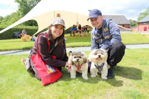 Josh Smith A man and a brunette woman, with two white dogs between them. All four are wearing caps and are crocuhed down on a patch of grass. The couple are smiling and looking at the camera. Behind them is a white marquee with more people sat underneath it.
