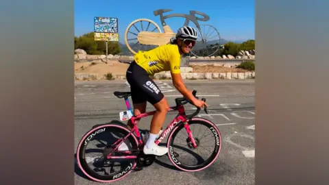 Michelle Sharland during the Tour de France Femmes. Michelle is wearing white cleets, black shorts and a yellow cycling top. She is on a pink bike with black wheels. She is wearing a silver helmet and is cycling in front of a giant silver bike statue.