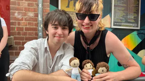 On the left, a young man with brown straight hair wearing a striped white shirt. On the right is Vex, who has a part-bleached mullet, wears sunglasses, a black necktie, and mesh black vest.