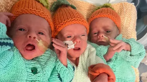 Ace & Shannon Page The trio of triplets lie together on a beige-coloured blanket. They are wearing matching green cardigans and knitted orange hats. All three are lying on their backs and have their eyes and mouths open