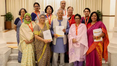 Nayem Ali/ CA Press Wing A group of 11 people stand in a marble hallway. All but two are women. The women wear bright saris, and appear to be a range of ages. In the middle is Yunus, the interim leader, wearing a blue and grey suit. He and two of the women hold reports in their hands.