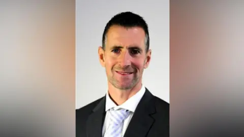 Wayne Briggs wears a suit and tie, he looks at the camera