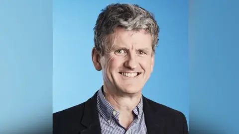 Cambridge University Hospitals NHS Foundation Trust Photo of Professor Miles Parkes against a blue background. He is wearing a back jacket and blue check shirt. He is smiling at the camera.