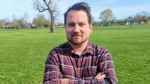 Contributed William Patrick is standing in a field, with trees and an Oak tree in the background. He is wearing a plaid red pink and maroon shirt. He has a beard and has his arms folded in front of him. 