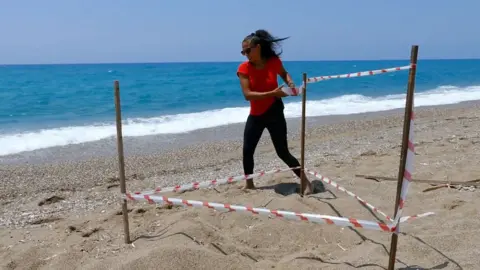 Tread the Globe A woman on a beech is wrapping plastic tape around three wooden poles which are arranged in a triangular shape, behind her the sea is visible