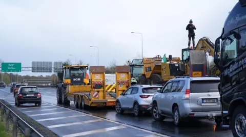 PA Media Fuel protesters block a motorway near Dublin - there are JCB trucks and diggers. One protesters is standing on a digger. There are green road signs.