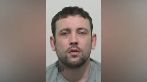 Mugshot of Wilkinson. He has a short dark beard and short black hair, with a tattoo on his neck.