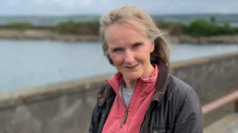 A woman with greying blonde hair in a dark jacket with a pink fleece underneath stands in front of Stranraer harbour