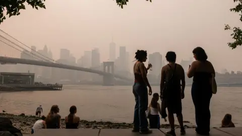 Spencer Platt / Getty Images People are silhouetted against the New York City skyline with Brooklyn bridge and the river in view, and a hazy smokey sky, in Brooklyn on 16 July.