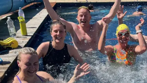 Four swimmers, a mixture of men and women, who are taking part in the challenge, are in the pool posing for the camera. For most, their hands are in the air and they are smiling.