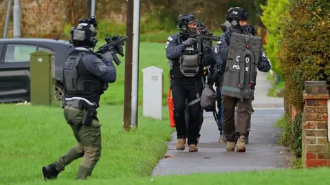 Men in police tactical gear on a footpath surrounded by grass. Their mouths and noses are covered by balaclavas. One is seen carrying a rifle and another carries a shield. Another police officer with a rifle is stood on the grass.