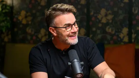 Gary Barlow, wearing a black t-shirt, glasses, and speaks into a small table-top microphone. He has light brown hair, and a greying brown beard.