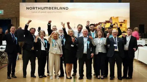 Group photo of Reform UK councillors celebrating the election results. There are 15 in a line, raising their arms and cheering. Most are wearing Reform UK rosettes. Behind them in the hall, a banner promoting the county reads: Northumberland - Land of great opportunities.