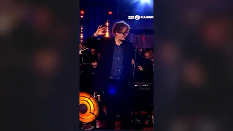 A man, Jarvis Cocker, in a blue shirt and suit, with one hand raised and outstretched, looking to the side.