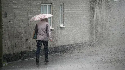 Getty Images A pistillate   walking distant  from the camera successful  dense  rain. She is wearing a grey jacket, with a container  implicit    her near  shoulder. She is carrying a floral umbrella. 