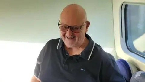 Wythenshawe Community Rugby Club Barry smiles wearing a black polo shirt while sat on public transport, with sun shining through a window. 