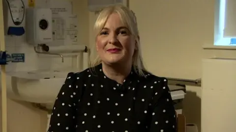 A woman with blonde shoulder-length hair looks at the camera. She is wearing a black top with white spots on it and behind her is a medical room which has a sink and tap and part of a window. 