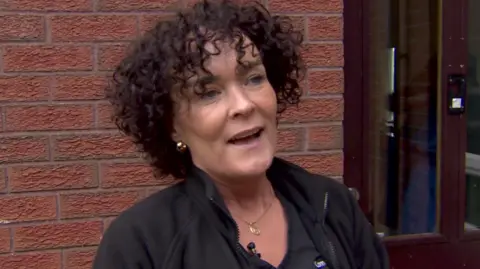 Bronagh has short black curly hair. She is wearing a black top and fleece. She is standing outside against a brick wall beside a door.