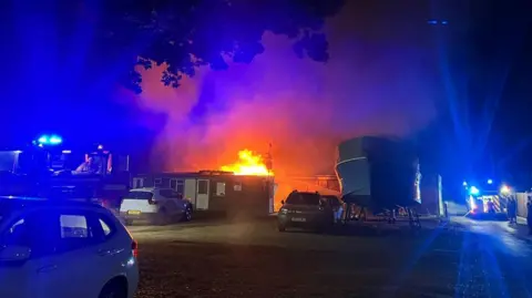 Contributed Flames shoot out of a building in the distance next to a large boat, taken by a photographer in a car park at night time.