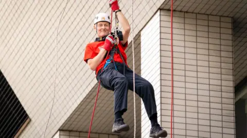 Nigel Haselden dangling from his abseiling rope as he descends from the hospital, he is wearing a red T-shirt, dark coloured trousers, a white safety helmet and red gloves