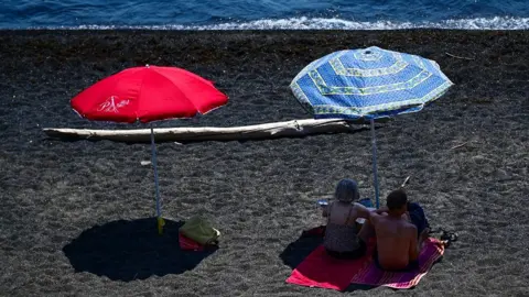 A red and blue parasol on a beach next to the sea, which is at the top of the picture. Two beachgoers are resting in the shade under the blue parasol on the right.