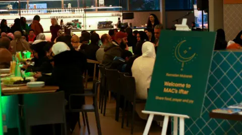 Midlands Arts Centre Rows of people wearing religious clothing sat down and eating. A green sign says Welcome to Mac's Iftar.