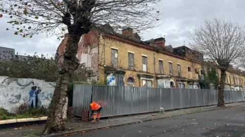 A man in orange hi-vis workwear is standing and inspecting a metal fence in front of a row of boarded up terraced houses. At the end of the terrace there appear to be brick remnants of a property no longer standing. A wall next to the man has a mural with some people standing together on it. There are two trees in view.