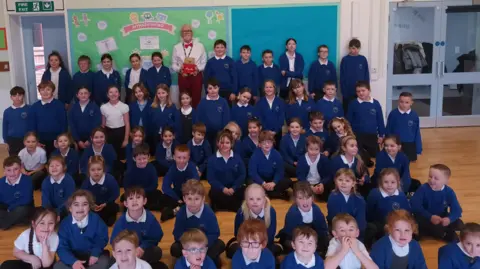 A mixed age range and gender group of pupils at Hertford Vale Primary School sit in a school hall. They wear blue school uniform jumpers and white polo shirts. Bob Hall, wearing a white shirt, red trousers and a red bow tie, stands at the back holding a teddy bear toy.