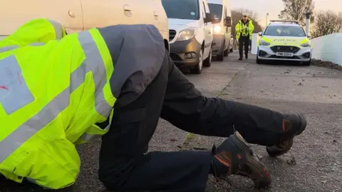 A man in a high-vis, yellow coloured jacket kneels on the floor next to car and looks at its tyres. Other white cars line up behind it. A police car is parked next to them. A police officer in uniform walks past.