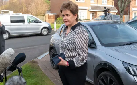 Nicola Sturgeon has short brown hair and is wearing a black and white top and black trousers. She is holding a black purse and is standing on the driveway of her house. There is a grey car in the driveway.