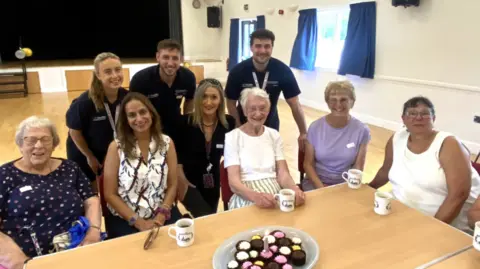 A group of elderly women are sat at a table drinking tea with members of the Worcestershire County Council's Public Health Team. They are sat in a hall setting. 