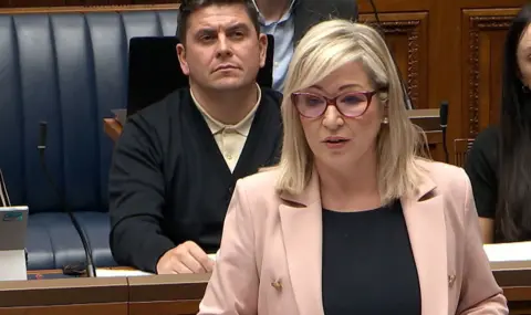 Michelle O'Neill in the assembly on monday. she has a pink jacket and blonde hair. She wears read winged glasses