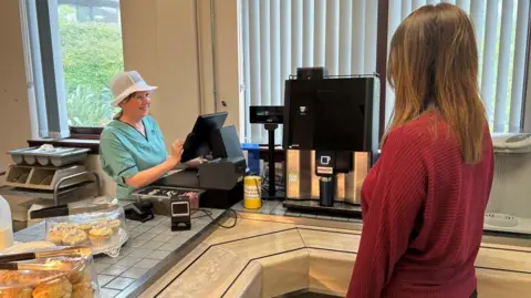 A female member of staff with long brown hair and wearing a red sweater is at the counter of an NHS cafe. There is a member of the cafe's staff at the till and some scones and cakes on display on the counter top. There is also a hot drinks machines with a reusable takeaway cup being used. 