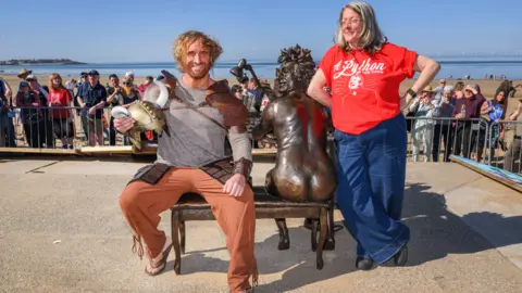 BBC Crowd attends unveiling ceremony on a beach as two people pose for a photograph in front of statue of naked Terry Jones playing a piano