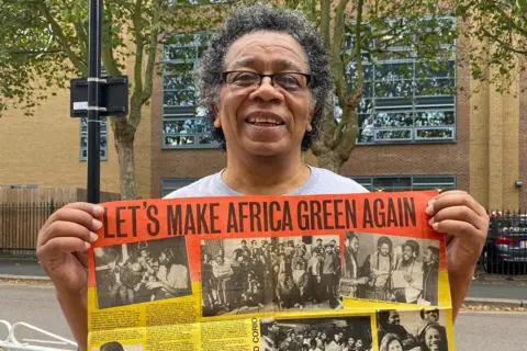 Hackney Council Leon Leiffer wearing glasses and a grey T-shirt holding up a newspaper page with Let's Make Africa Green Again written across the top