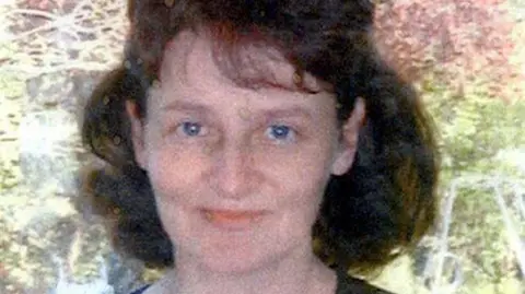 Wiltshire Police A grainy photograph of a woman with shoulder-length brown hair. She is wearing a black scoop-neck top and is standing in front of a tree smiling.