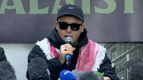 A man is talking into a microphone. He's dressed in a black hoodie with a red and white scarf on his shoulders. 