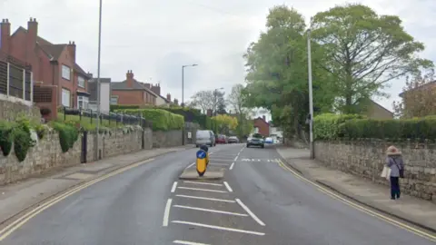 Google An A-road with semi-detached homes on either side. A woman walks along the pavement on the right. In the middle of the road is a crossing.