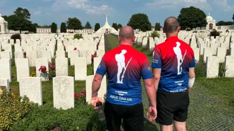 RBVE Two men wearing red and blue cycling gear. They are facing a row of graves inside a war cemetery.
