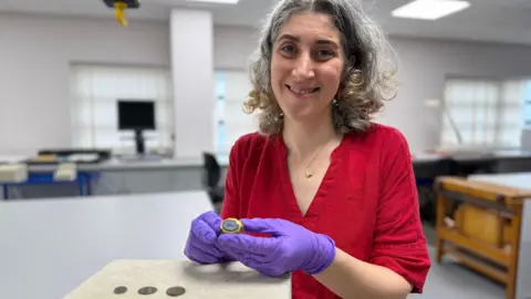 Woman wearing a red top holding the gold Roman ring with purple gloves on. There are some coins in the bottom left of the picture too.