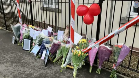 Flowers, cards and pictures left on a pavement outside a set of railings.