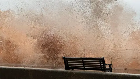 Sarah H/BBC Weather Watchers This picture shows a large wave crashing over a sea wall or promenade, sending a huge spray of water into the air. In the foreground, there is a black bench positioned on the walkway, which appears to be very close to the impact zone of the wave. The water looks turbulent and muddy.