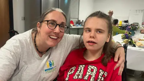 A woman in a grey top has her hand around a girl with long brown hair, wearing a red t-shirt. The woman has black glasses and long brown hair. They both smile as they look of-camera to the right. Behind them is a white-walled kitchen with a grey fridge in the middle. 